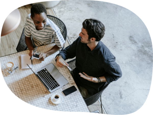  Two coworkers sitting at a table with a laptop, notebook, and coffee, engaging in a discussion in a casual workspace.