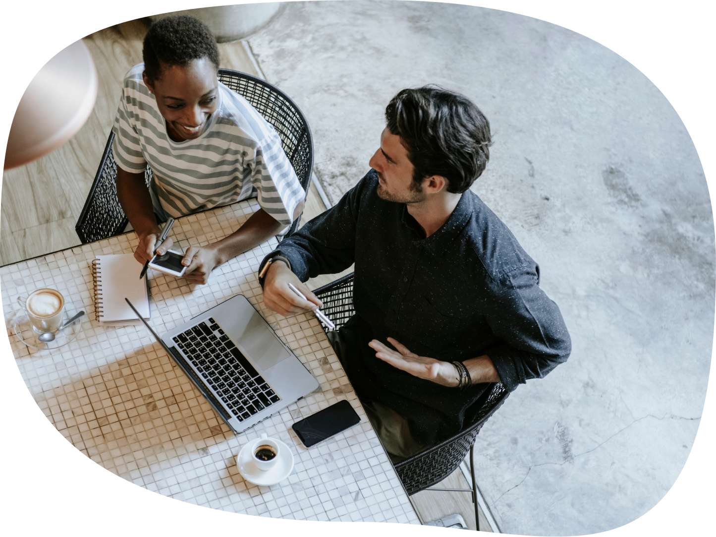 Two coworkers sitting at a table with a laptop, notebook, and coffee, engaging in a discussion in a casual workspace.