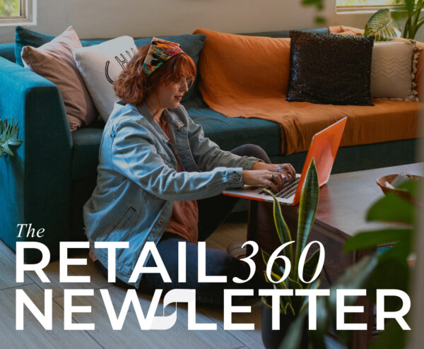 A young woman with curly red hair, wearing a denim jacket and a colorful headband, sits on the floor in a cozy living room, working on an orange laptop placed on a wooden coffee table. The room features a teal sofa with decorative pillows and an orange throw blanket. Green plants add a natural touch to the space. Overlaying the image is the text 'The RETAIL 360 NEWSLETTER' in bold, modern typography.